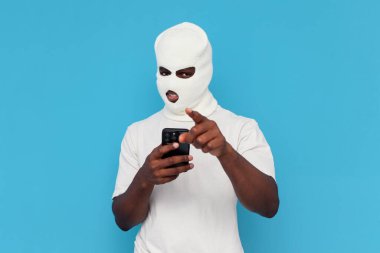 Seni seçiyorum. Beyaz kar maskeli Afrika kökenli Amerikalı erkek hacker mavi arka planda akıllı telefon kullanıyor, maskeli zorba internette suç işliyor ve tehdit ediyor.
