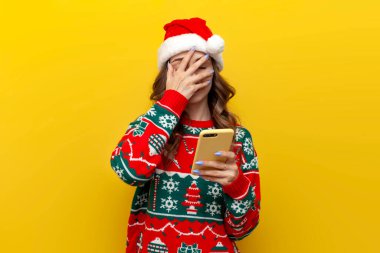 Şaşkın kız yılbaşı süveteri ve Noel Baba şapkası takıyor. Akıllı telefon kullanıyor ve sarı izole arka planda facepalm gösteriyor. Telefonunu tutan kadın yeni yıl kıyafetleriyle elini yüzünü kapatıyor.