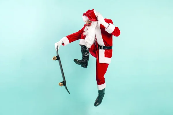サングラスとスーツのサンタクロースは 青い隔離された背景にスケートボードでジャンプしてハエ ヒップスターサンタはスケートボードに乗り 新年とクリスマスのために空気をトリックします — ストック写真