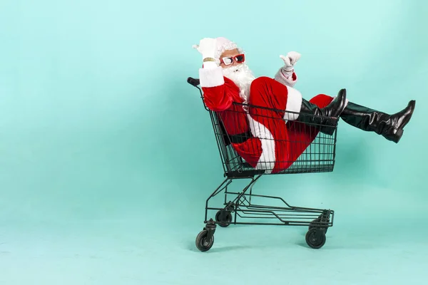 サンタクロースの買い物客は空のショッピングカートに座り 青い背景を指し サンタは新年のショッピングのためにスーパーマーケットに急いで クリスマスと背後からコピースペースを指します — ストック写真