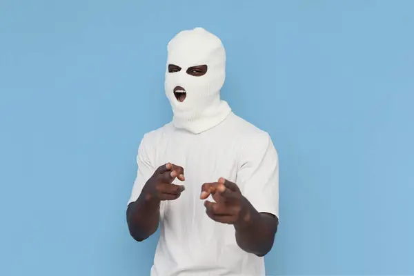 I choose you. african american male bully in white balaclava threatens and points with his hands at the camera on blue isolated background, unrecognizable guy in mask screams