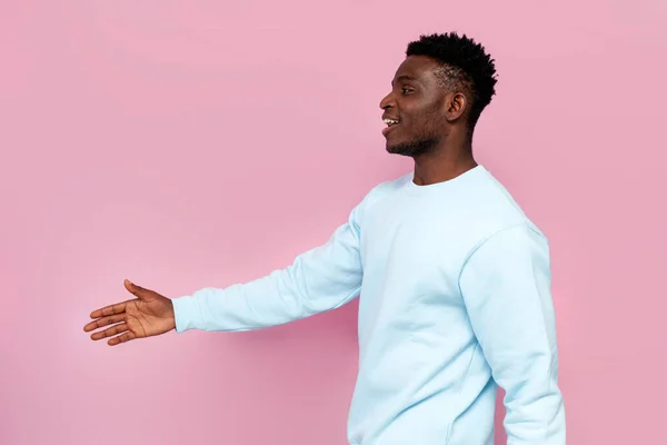 stock image african american man in blue sweater smiles and holds out his hand for handshake on pink isolated background, side view