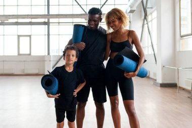 Afrika kökenli Amerikalı genç aile spor salonunda bir arada duruyor ve yoga minderi tutuyorlar. Baba ve oğul spor kıyafetleriyle spor salonunda karemat eğitimindeler.