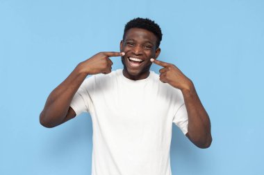 Beyaz tişörtlü Afro-Amerikan adam gülümsüyor ve mavi izole arka plandaki dişlere işaret ediyor, adam dişlek gülümseme reklamları yapıyor.