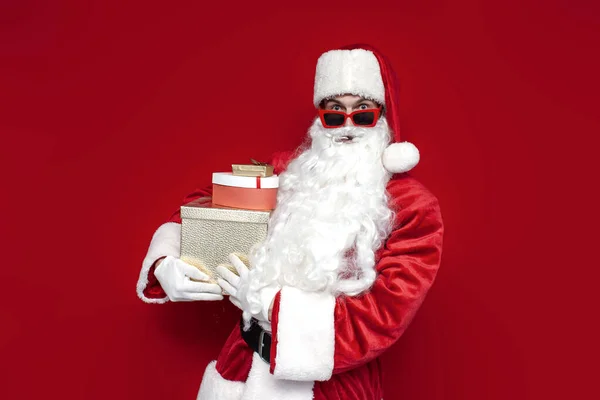 shocked santa claus in christmas suit and glasses holds boxes with gifts and is surprised on red background