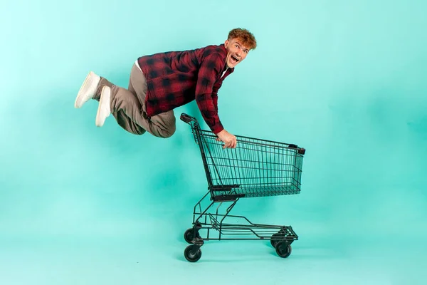 young crazy guy shopper jumps and rejoices with shopping cart from supermarket on blue isolated background, male customer quickly runs in the air for shopping