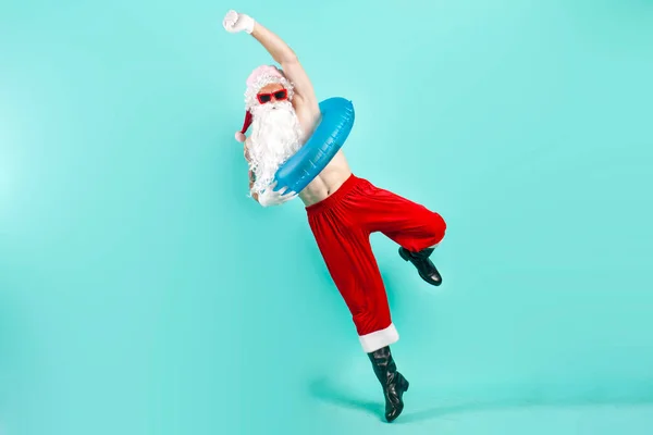 santa claus in suit with inflatable swim ring in sunglasses jumps and flies and rejoices in success on blue isolated background, santa tourist rides on vacation at the sea and celebrates victory