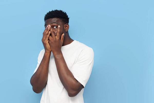 frightened young guy african american in white t-shirt covers his face with his hands and peeps on blue isolated background, modest man is shy and looks out at copy space