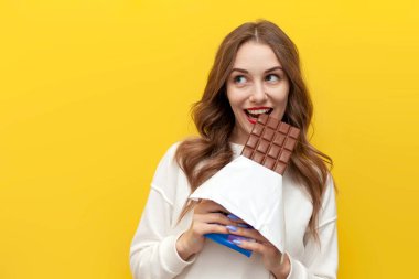 Genç kız büyük çikolata yiyor ve ısırıyor ve sarı izole edilmiş arka planda hayaller kuruyor, tatlı yemekleri olan mutlu kadın fotokopi alanına bakıyor.