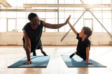 Baba oğul spor yapıyor ve babasıyla birlikte beş dakika beşlik çakıyor, Afrikalı Amerikalı çocuk yoga minderinde yerden şınav çekiyor ve başarı jesti yapıyor.