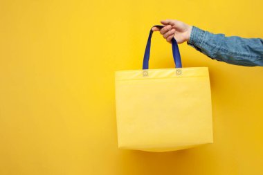 Sarı arka planda boş bir kumaş torba, adamın elinde yeniden kullanılabilir plastik olmayan bir çanta, plastik ve ekolojiyi reddetme konsepti