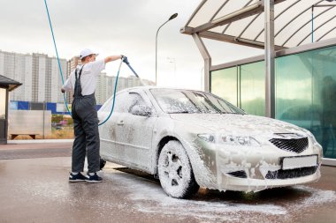 Üniformalı genç adam araba yıkama işçisi arabayı hortumla kaplıyor, tulumlu adam dışarıda araba temizleme servisinde çalışıyor.