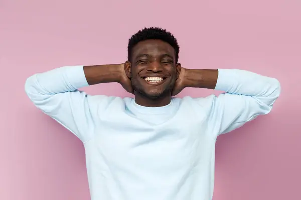 african american man in blue sweater rests and dreams with his eyes closed on pink isolated background, man lies and relaxes
