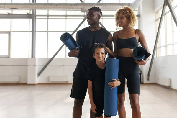african american family in sportswear with yoga mat stands in the gym and smiles, mom dad and son on fitness workout
