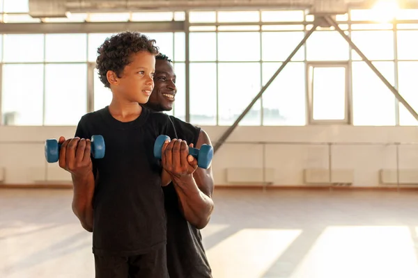 father and son go in for sports in the gym, african american man helps his son lift dumbbells and teaches healthy lifestyle, the boy trains with dad and looks at copy space