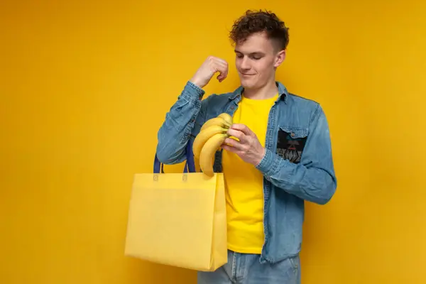 young guy a buyer with a fabric eco bag buys bananas and puts them in a reusable non-plastic bag on a yellow background, ecology concept
