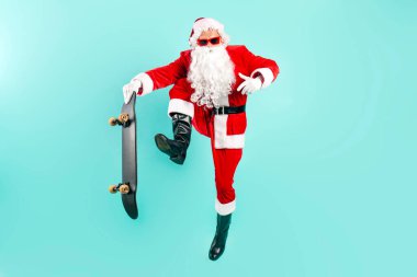 Güneş gözlüklü ve takım elbiseli Noel Baba mavi arka planda kaykayla zıplar ve uçar. Hipster Noel Baba kaykaya biner ve yılbaşı ve yılbaşı için havada numara yapar.