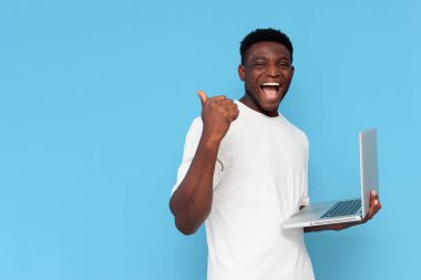 Beyaz tişörtlü Afro-Amerikan adam dizüstü bilgisayar kullanıyor ve mavi izole arka planda gösteriyor. Kazanan kişi online başarıya seviniyor ve reklam boşluğunu arkadan kopyalıyor.