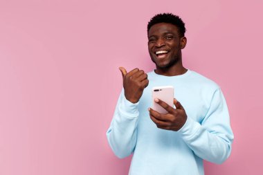 Mavi kazaklı Afro-Amerikan adam akıllı telefon kullanıyor ve pembe izole arka plandaki boş alanı işaret ediyor.