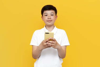 Küçük Asyalı çocuk sarı arka planda akıllı telefon kullanıyor, Koreli çocuk izole edilmiş arka planda cep telefonunda çevrimiçi yazıyor.