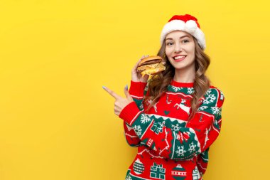 Noel süveteri ve Noel Baba şapkası giyen genç tatlı bir kız büyük çizburger yiyor ve sarı izole arka planda boş bir alana işaret ediyor, kadın yeni yıl için fast food yiyor ve uzay reklamları yapıyor.