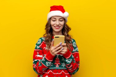 Noel süveteri ve Noel Baba şapkalı genç kız sarı izole edilmiş arka planda akıllı telefon kullanıyor, kadın telefonda noel kıyafetleri içinde mesaj yazıyor.