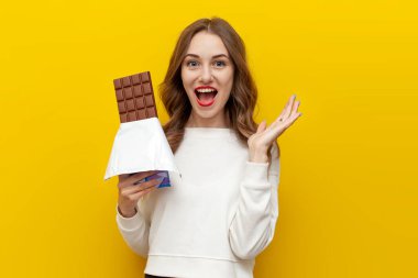 Genç kız büyük çikolata çubukları tutuyor ve sarı izole edilmiş arka planda çığlıklar atıyor ve seviniyor. Tatlı yemekleri olan şok olmuş kadın şaşırıyor ve şaşırıyor.