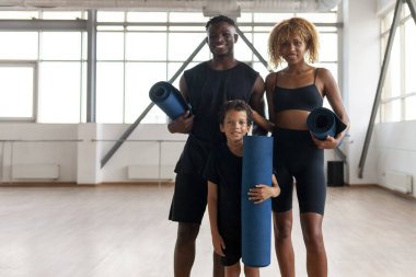 Afrika kökenli Amerikalı genç aile spor salonunda bir arada duruyor ve yoga minderi tutuyorlar. Baba ve oğul spor kıyafetleriyle spor salonunda karemat eğitimindeler.