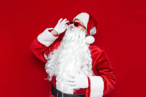 赤い眼鏡のサンタクロースは 色の背景 帽子とサンタのコスチュームで彼の口ひげに触れます クリスマス — ストック写真