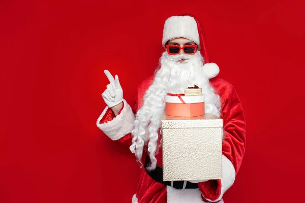 クリスマスのコスチュームとメガネのサンタクロースは 赤い背景のコピースペースに彼の手で贈り物とショーを持つボックスを保持しています — ストック写真