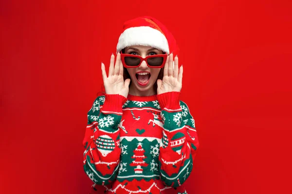 身穿圣诞毛衣 头戴圣诞礼帽的可爱女孩看着眼镜 红着脸大喊 震惊的圣诞女扮男装表现出惊喜 — 图库照片