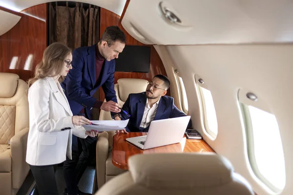 business people at meeting in private luxury jet, campaign workers in suits discussing project and looking at laptop on the flight, business partners communicate and work with documents on the plane