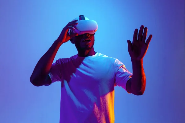 stock image african american male gamer in virtual reality glasses touches his hands in neon lighting, the guy uses modern vr gadget and chooses and points his finger at copy space