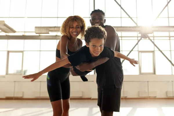 african american family in sportswear holding and carrying son in the air and playing in gym, boy in the arms of his parents is flying and having fun, the concept of freedom and healthy lifestyle