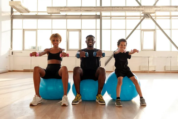 sporty african american family is doing pilates on fitness ball in the gym, boy with his parents is holding dumbbells and doing exercises, mom dad and son are warming up