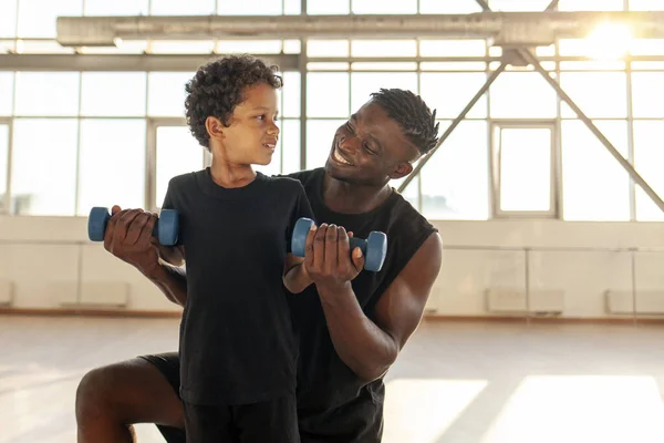 father and son go in for sports in the gym, african american man helps his son lift dumbbells and teaches healthy lifestyle, the boy trains with dad and looks at copy space