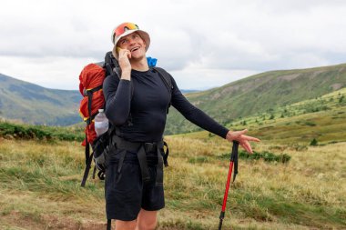 Sırt çantalı ve yürüyüş ekipmanlı bir erkek turist, dağların arka planında telefonla konuşuyor ve gülümsüyor, dağ yürüyüşündeki adam cep telefonuyla iletişim kuruyor.