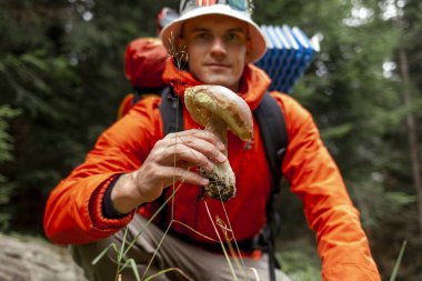 Genç adam ormanda beyaz mantar tutuyor, dağ yürüyüşünde turist mantar topluyor.