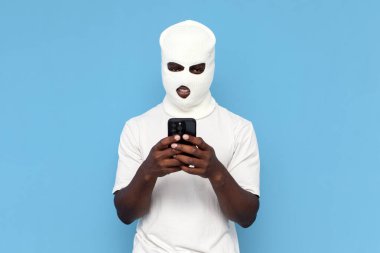 Beyaz kar maskeli Afrika kökenli Amerikalı erkek hacker mavi arka planda akıllı telefon kullanıyor, maskeli zorba telefonda online suça karıştı.
