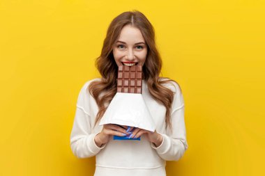 Genç kız büyük çikolata yiyor ve ısırıyor ve gülümsüyor ve sarı izole arka planda seviniyor, tatlı yemeği olan mutlu kadın dişlek bir gülümseme sergiliyor