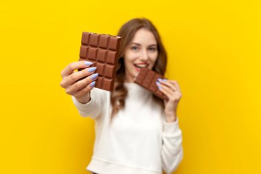 Genç kız büyük çikolata yiyor ve ısırıyor ve size sarı izole edilmiş arka planda bir parça sunuyor, mutlu kadın çikolata veriyor ve gösteriyor, beslenme ve diyet kavramı