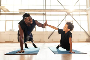 Afrikalı Amerikalı bir çocuk yoga minderi üzerinde egzersiz yapıyor ve babasıyla şınav çekiyor. Oğlunu eğiten ve beşlik çakan bir adam. Sağlıklı yaşam tarzı ve ebeveynlik kavramı.
