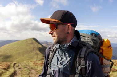 Ceketli ve güneş gözlüklü bir adam dağa çıkıp güneşe bakıyor. Dağ kıyafetleri ve ultraviyole gözlüklü bir turist.