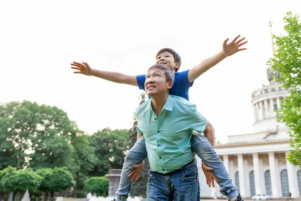 asian old father carries son on his back and they fly forward together in summer, korean boy play with grandpa outdoors