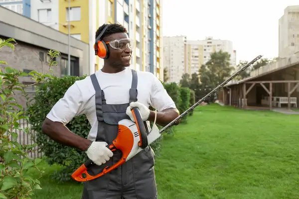 african american male garden worker in uniform trims bushes with electric tool, man in goggles and headphones works with brush cutter and trims trees and greenery