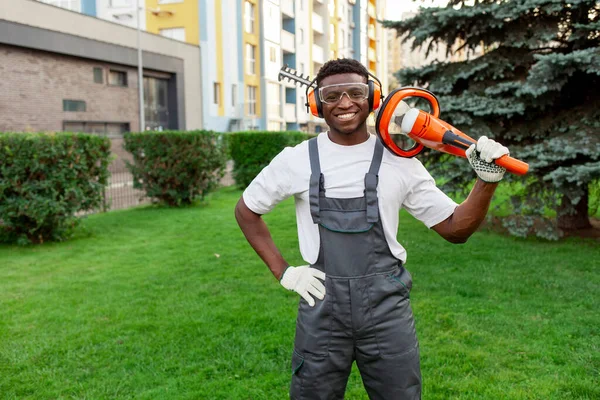 garden worker in uniform cuts bushes, african american man in goggles and headphones holds electric brush cutter and garden tool on background of greenery, copy space