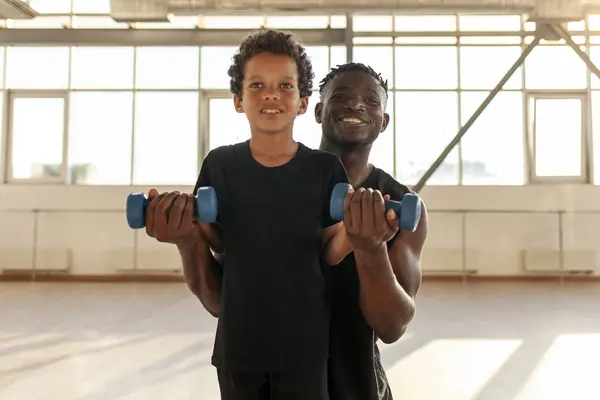 father and son go in for sports in the gym, african american man helps his son lift dumbbells and teaches a healthy lifestyle, the boy is exercising with dad