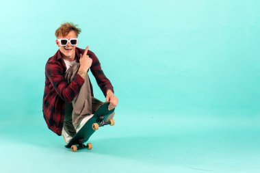 Genç neşeli adam mavi izole arka planda kaykaya biniyor, güneş gözlüklü hippi patenci ve reklam alanı kopyalıyor.