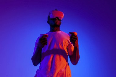 Sanal gerçeklik gözlüklü Afrikalı Amerikalı bir adam neon ışıkta joystickleri tutuyor, oyuncu gözlük kullanıyor ve kontrol panellerini tutuyor.