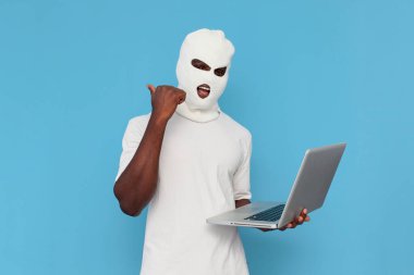 Beyaz kar maskeli Afro-Amerikan erkek hacker dizüstü bilgisayar kullanıyor ve mavi izole arka plana işaret ediyor, maskeli holigan çevrimiçi suça karıştı ve reklamlar arkadan uzayı kopyalıyor